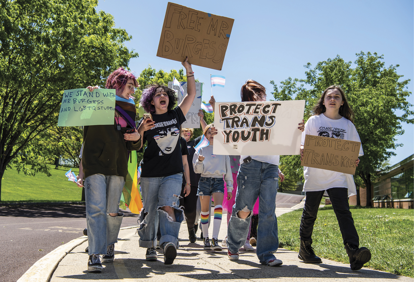 Students walking on sidewalk protesting with signs. One says