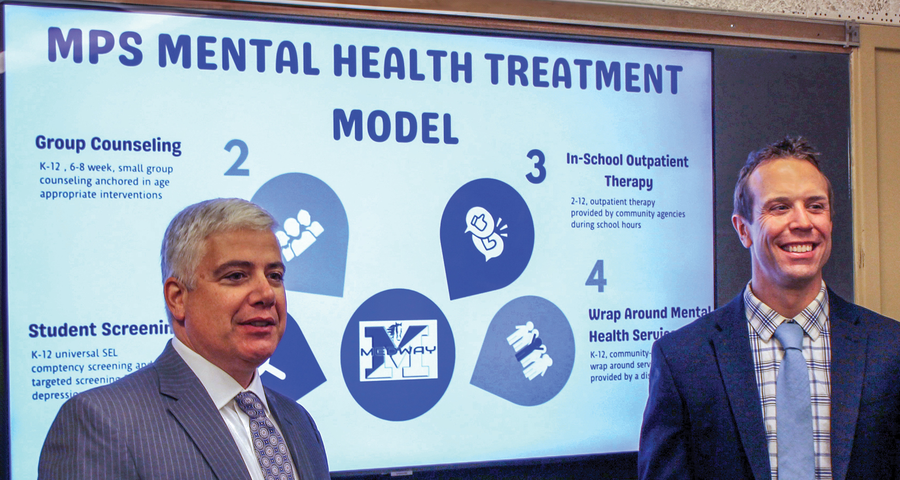 Two men in suits standing in front of a projection that reads,