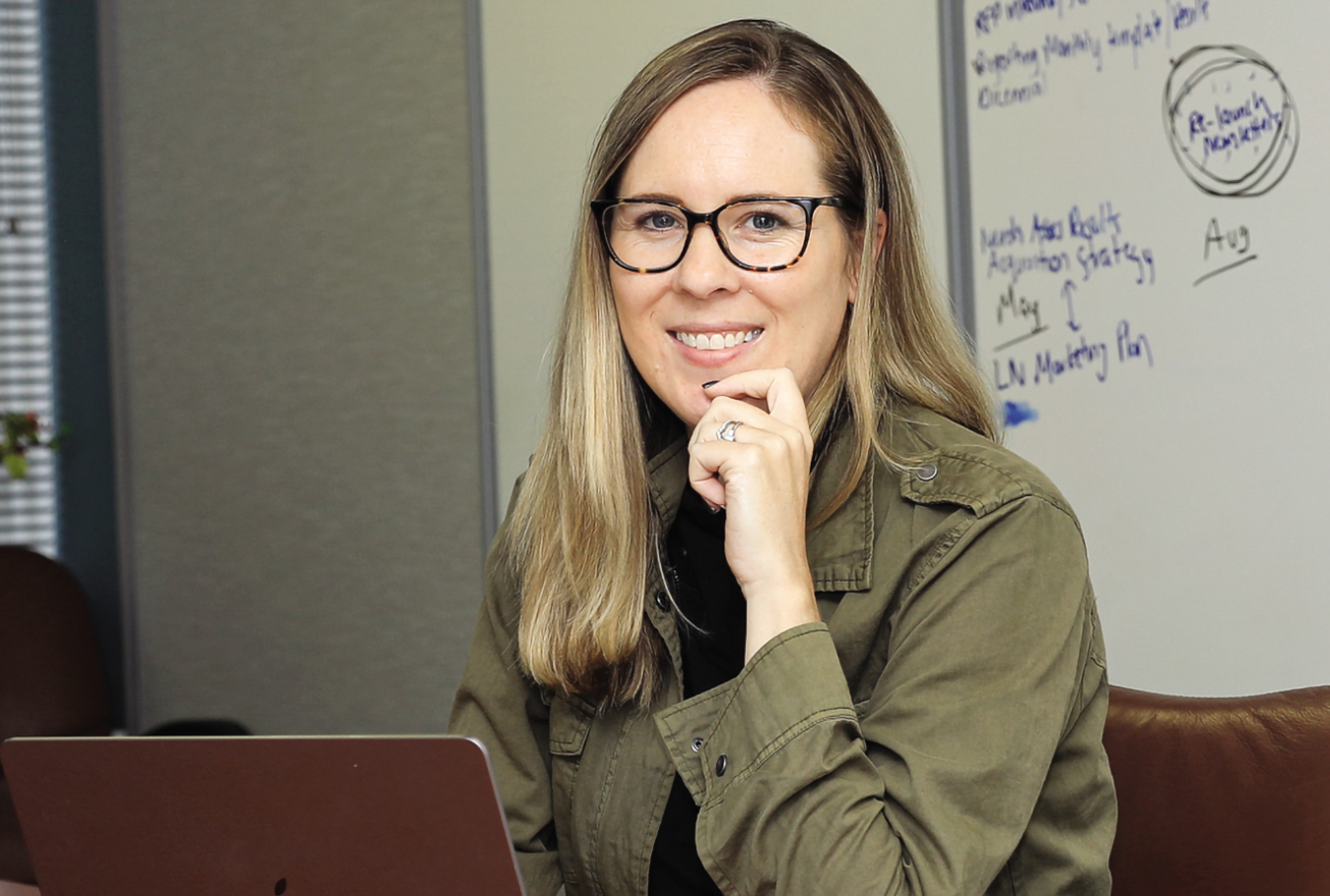 A blonde white woman wearing a green jacket sitting at her computer with black glasses