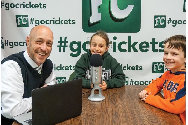 Joe Sanfelippo works on a podcast with two students in front of a banner with their twitter hashtag