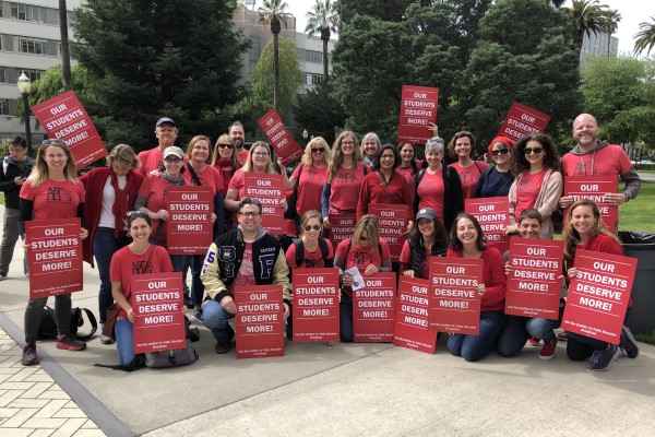A group of professionals stand with red signs reading Our students deserve more