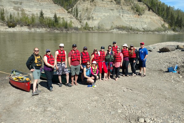 Several teachers stand on a beach in life jackets next to canoes