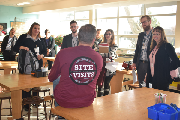 A group of educators in a classroom looking to the instructor wearing a shirt that says site visits