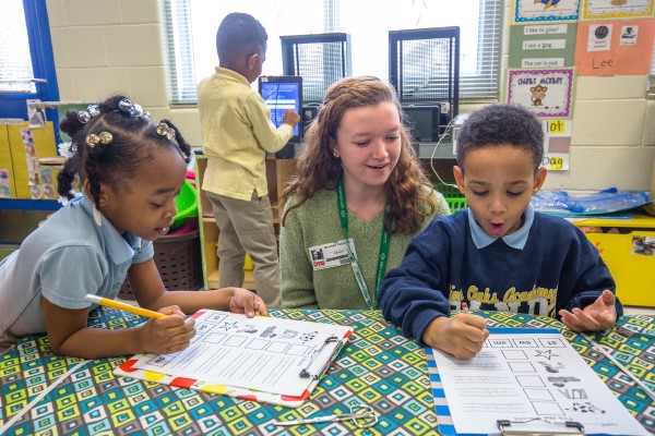 A woman assists two younger students with a worksheet in a classroom