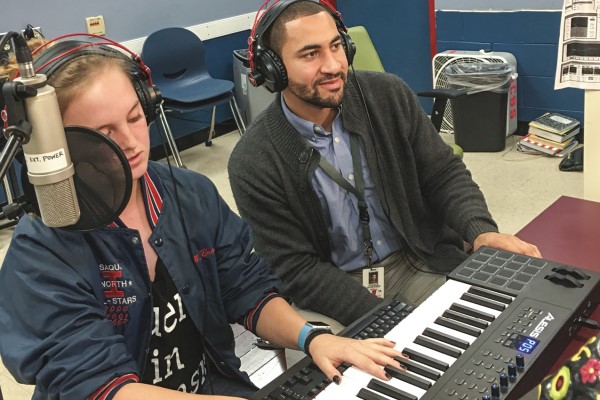 A student plays keyboard and sings while a teacher listens on headphones