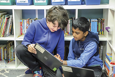 Two young boys show each other their laptops in a classroom