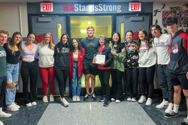 A large group of students stand together smiling at camera with their superintendent. "WAStandsStrong" is written above the doorway.