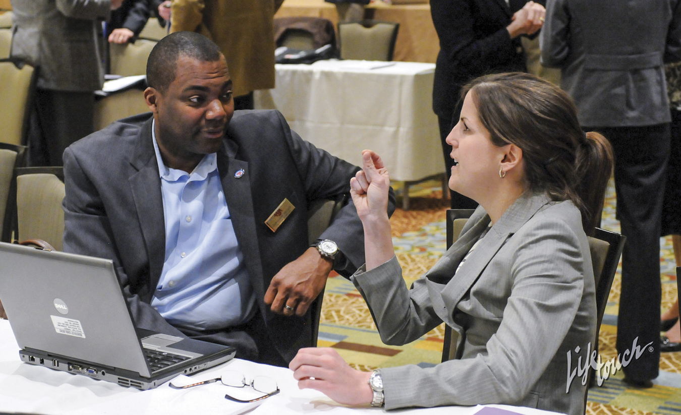 Black man in gray suit and blue button down talking to white woman with brown hair in light gray suit, both sitting at table with laptop