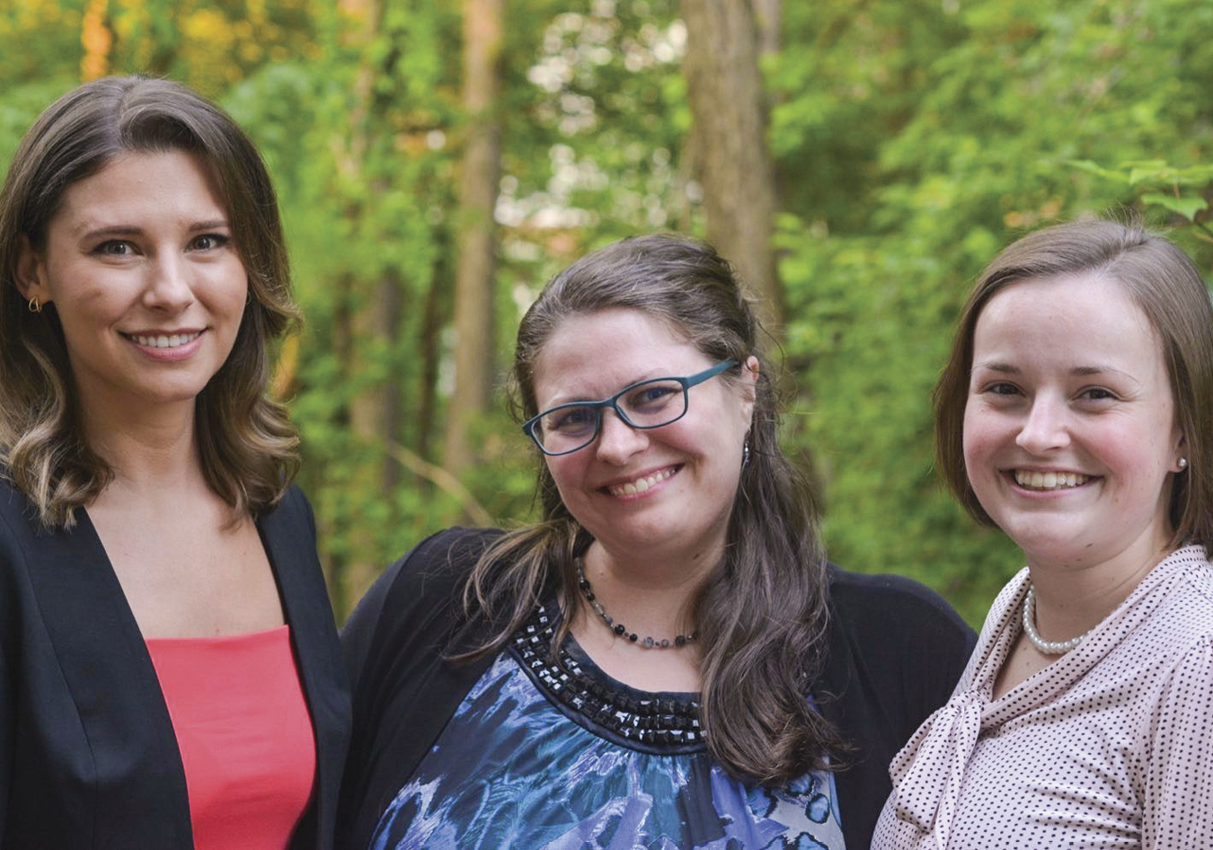 Three white women with brown hair smiling with forest behind them. Katherine is wearing a black blazer with salmon blouse, Amelia wears blue glasses and a blue top, Morgan wears a white blouse and pearl necklace