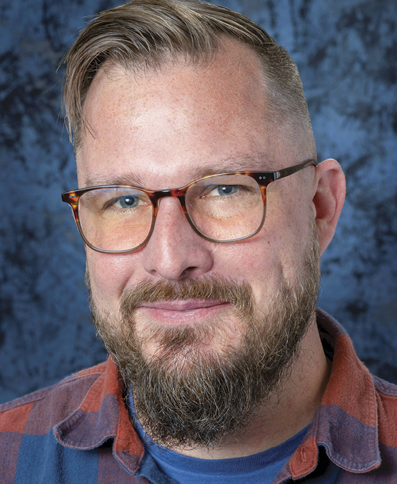 White man wearing checked shirt and brown glasses. He has a beard.