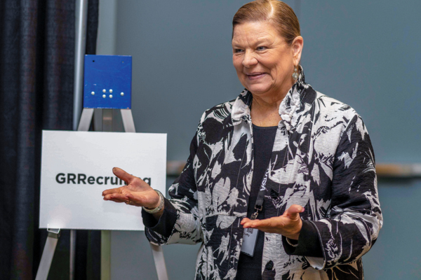 woman with brown hair gesturing wearing black and white shirt with sign behind her that says GRRecruiting