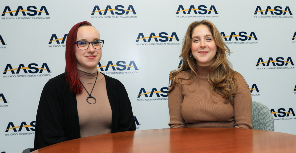 Two women sitting at a table in front of AASA backdrop. The woman on the left has pink hair on one side and a shaved head on the right side, wearing glasses and a black cardigan. The one on the right has honey brown hair and wearing a brown top.