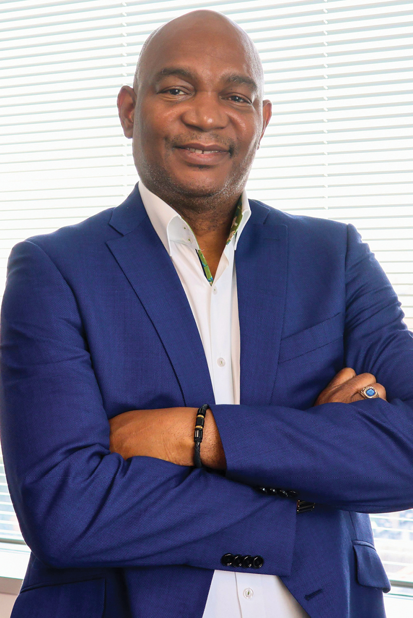 Olufemi Aina, a Black man, smiling with arms crossed in a white shirt and blue suit
