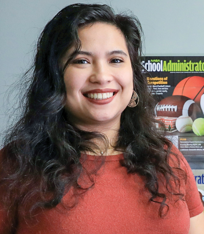 Jacqueline Hyman, a half Filipino half white woman with curly dark brown hair smiling wearing an orange top in front of a rack of School Administrator magazines