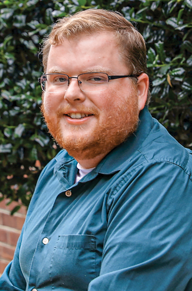 A white man with strawberry blonde hair and bear, wearing glasses and a blue button down shirt