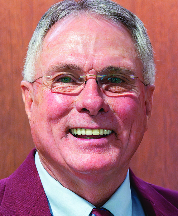 A headshot of Kim Alexander, a white man with gray hair and glasses, smiling wearing a red suit jacket