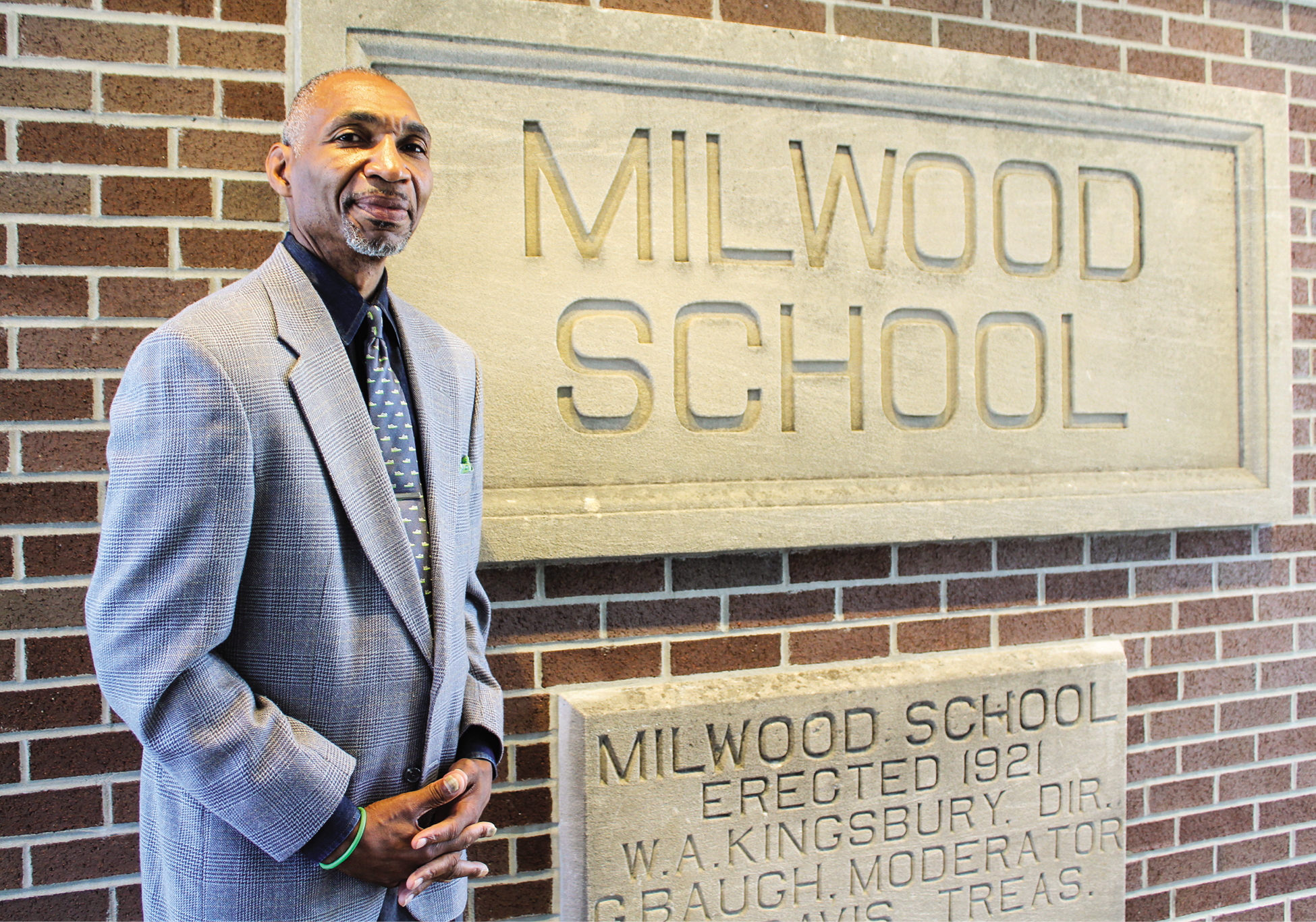 A bald Black man in a gray suit in front of a brick wall and sign that says Milwood School