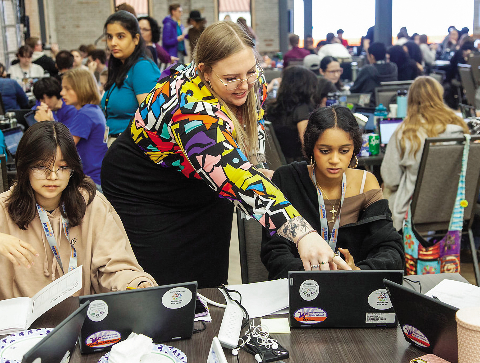 A white teacher pointing at the laptop screen of a young female student of color, surrounded by many other students and teachers