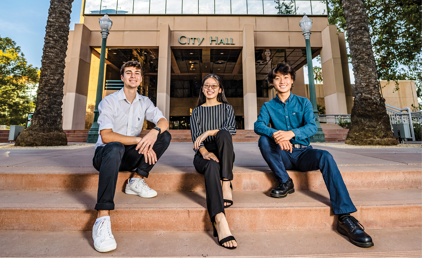 Three students sit on the steps in front of a City Hall building