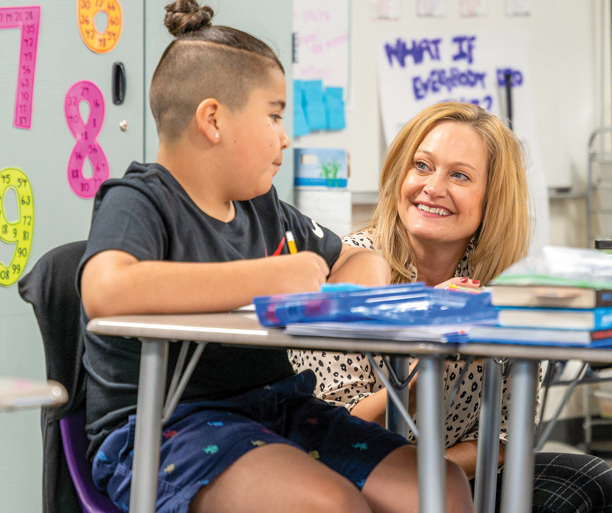 A woman kneeling next to a child's desk smiling