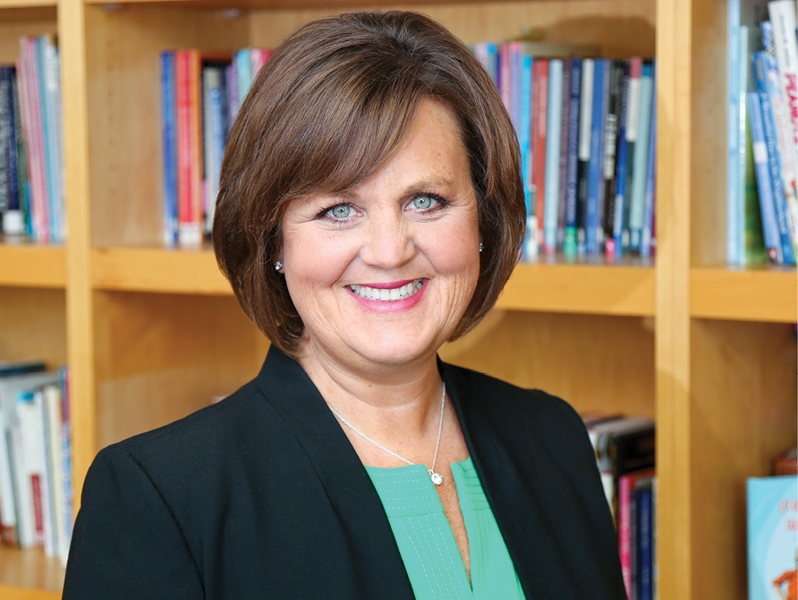 A headshot of a woman wearing a black suit and green top in front of a bookshelf