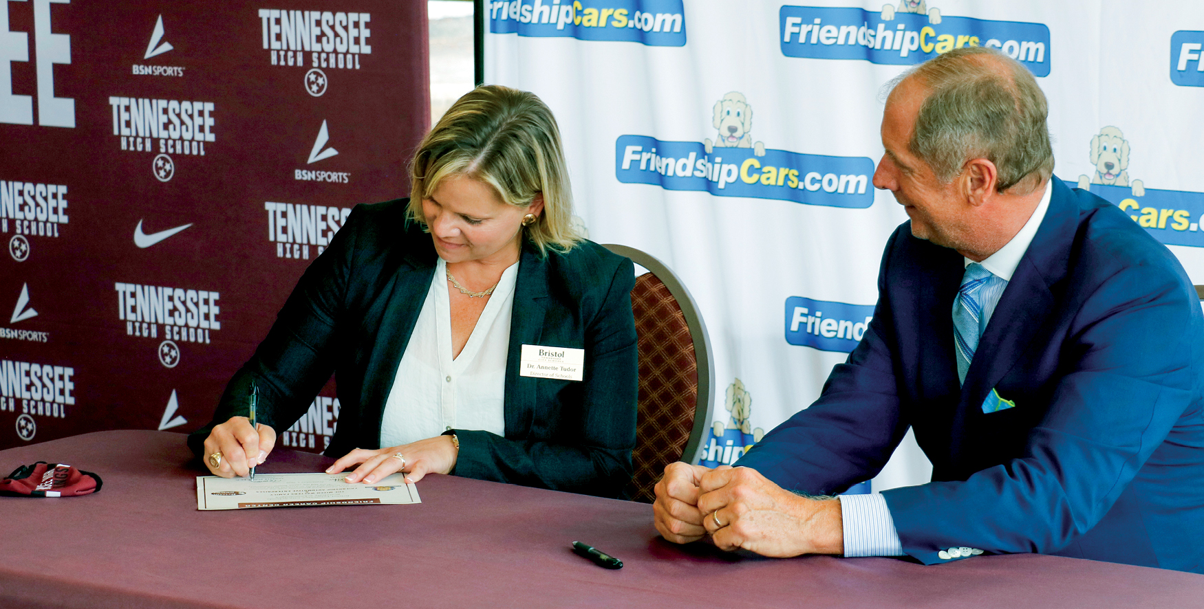 Blond woman in black blazer signs a document at table with a man in blue suit