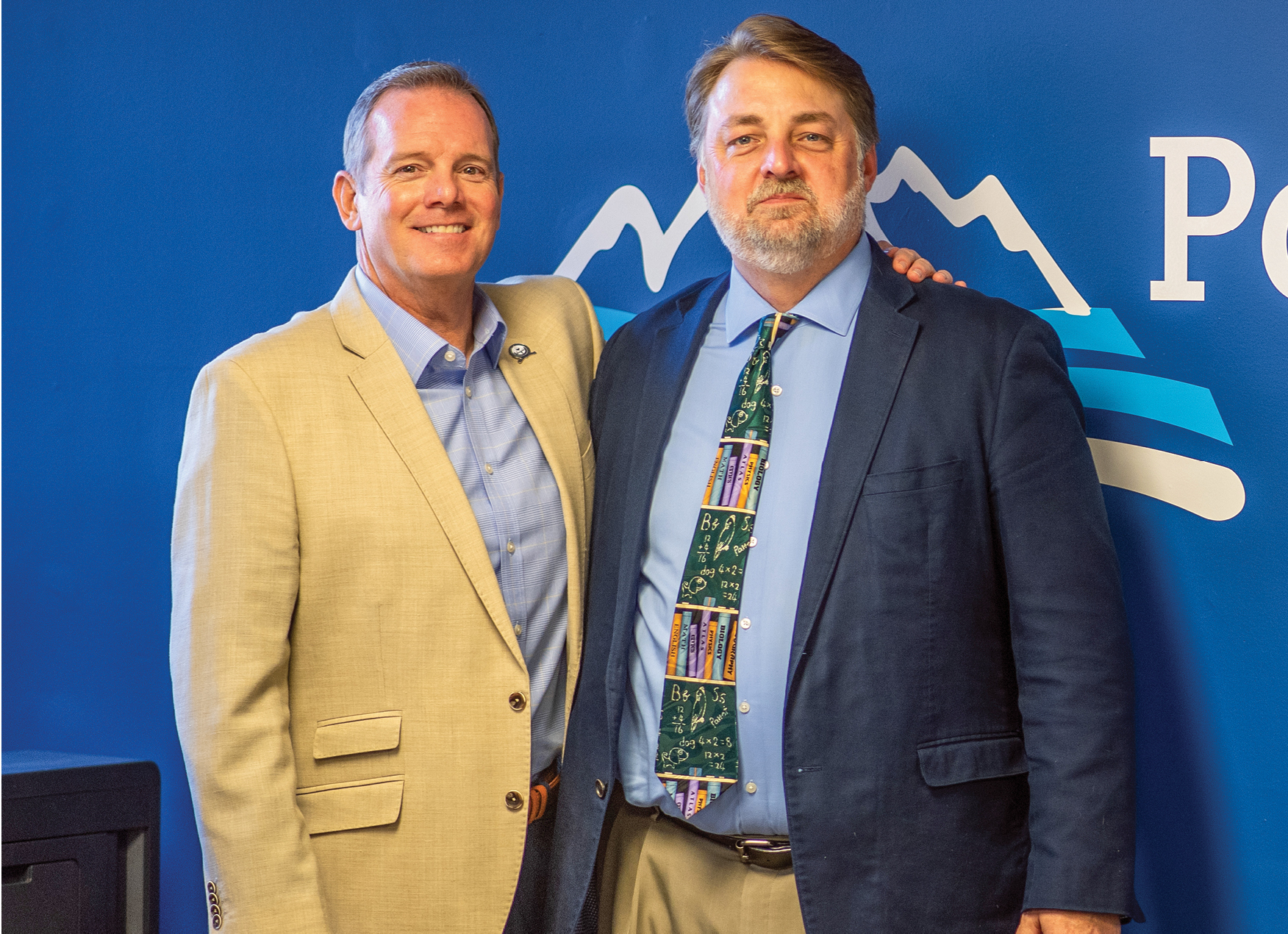 Two men in suits standing in front of school district logo on wall