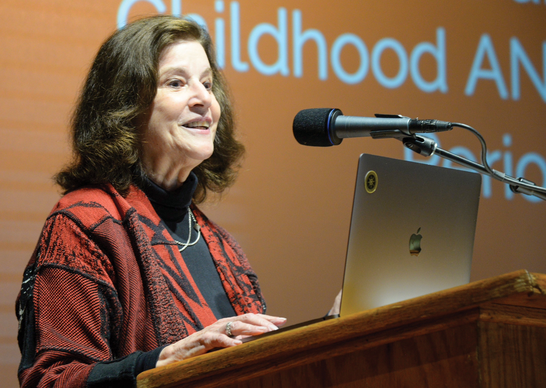 Woman with brown hair wearing red at podium with laptop and microphone