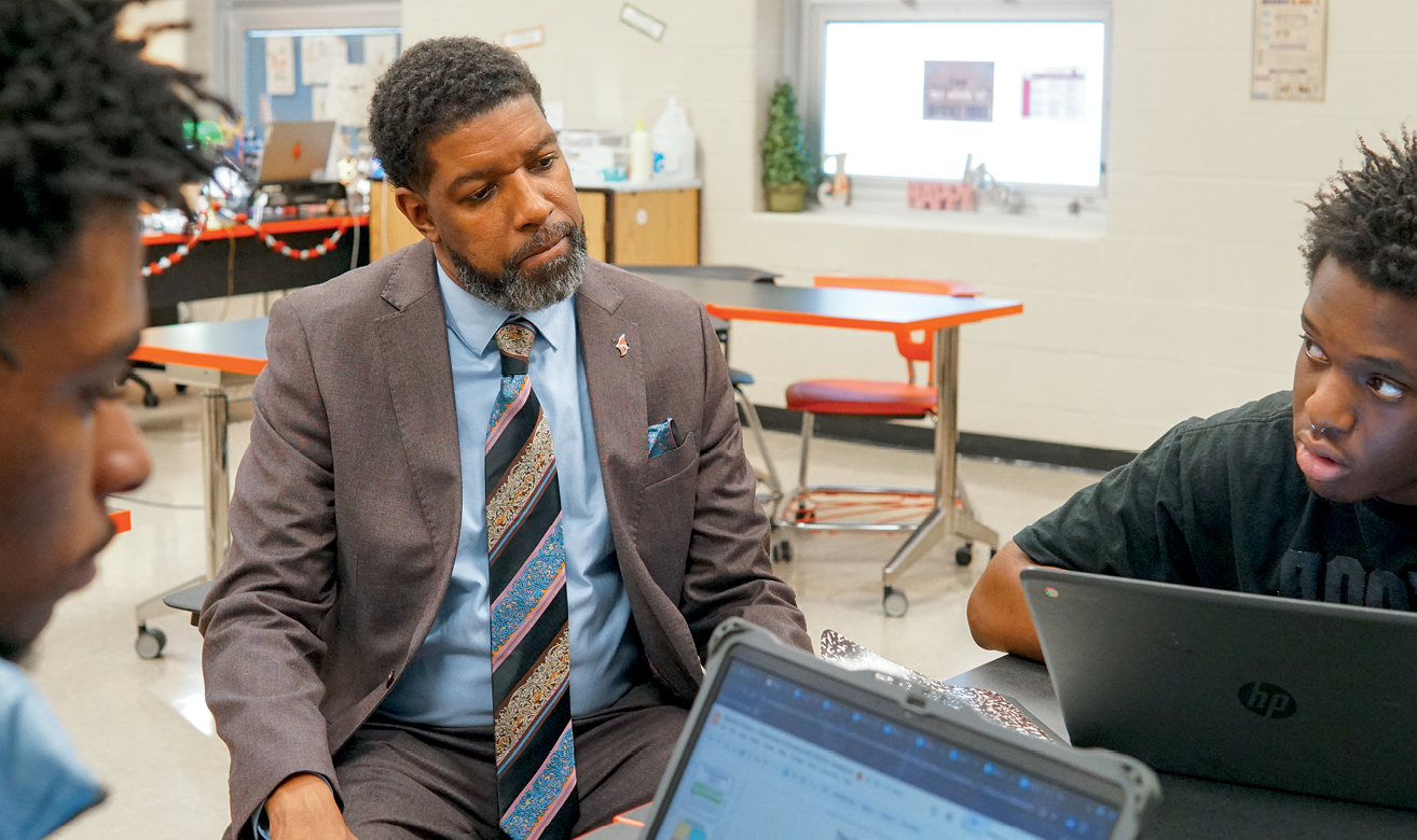 A Black man in suit sitting with two teenagers at table and laptops