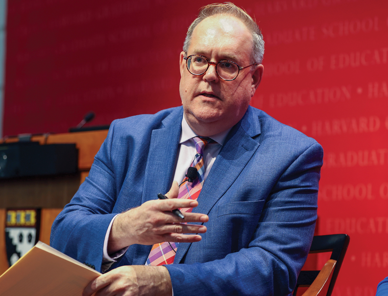 A man in blue suit with glasses talking and sitting