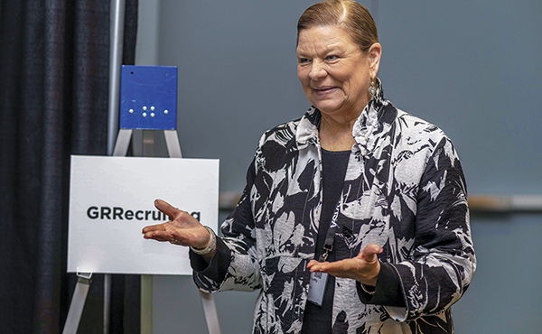 Woman talking with arms gesturing forward with sign behind her that says GRRecruiting