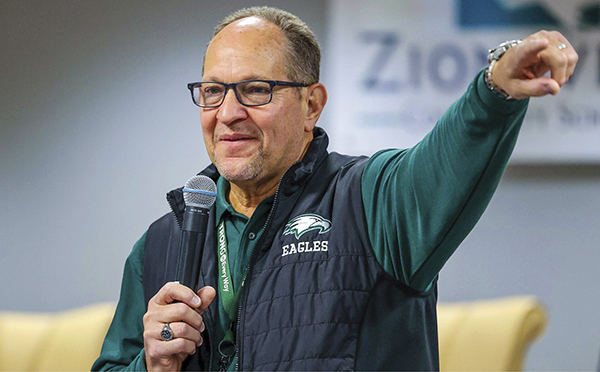Scott Robison with microphone pointing arm, wearing long sleeve green shirt with black vest and Eagles mascot